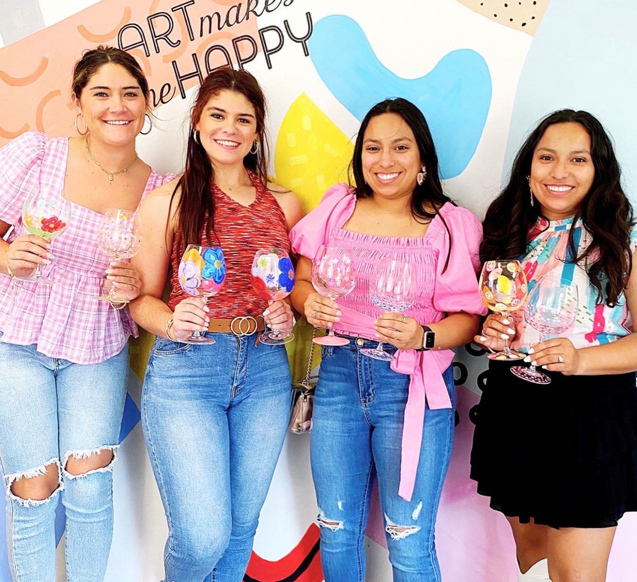 Four smiling women in casual jeans and bright tops at an indoor paint-and-sip workshop, each holding a colorful hand-painted wine glass in front of a vibrant abstract wall mural.
