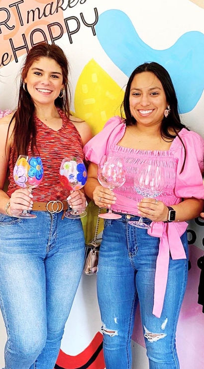 Four smiling women in casual jeans and bright tops at an indoor paint-and-sip workshop, each holding a colorful hand-painted wine glass in front of a vibrant abstract wall mural.