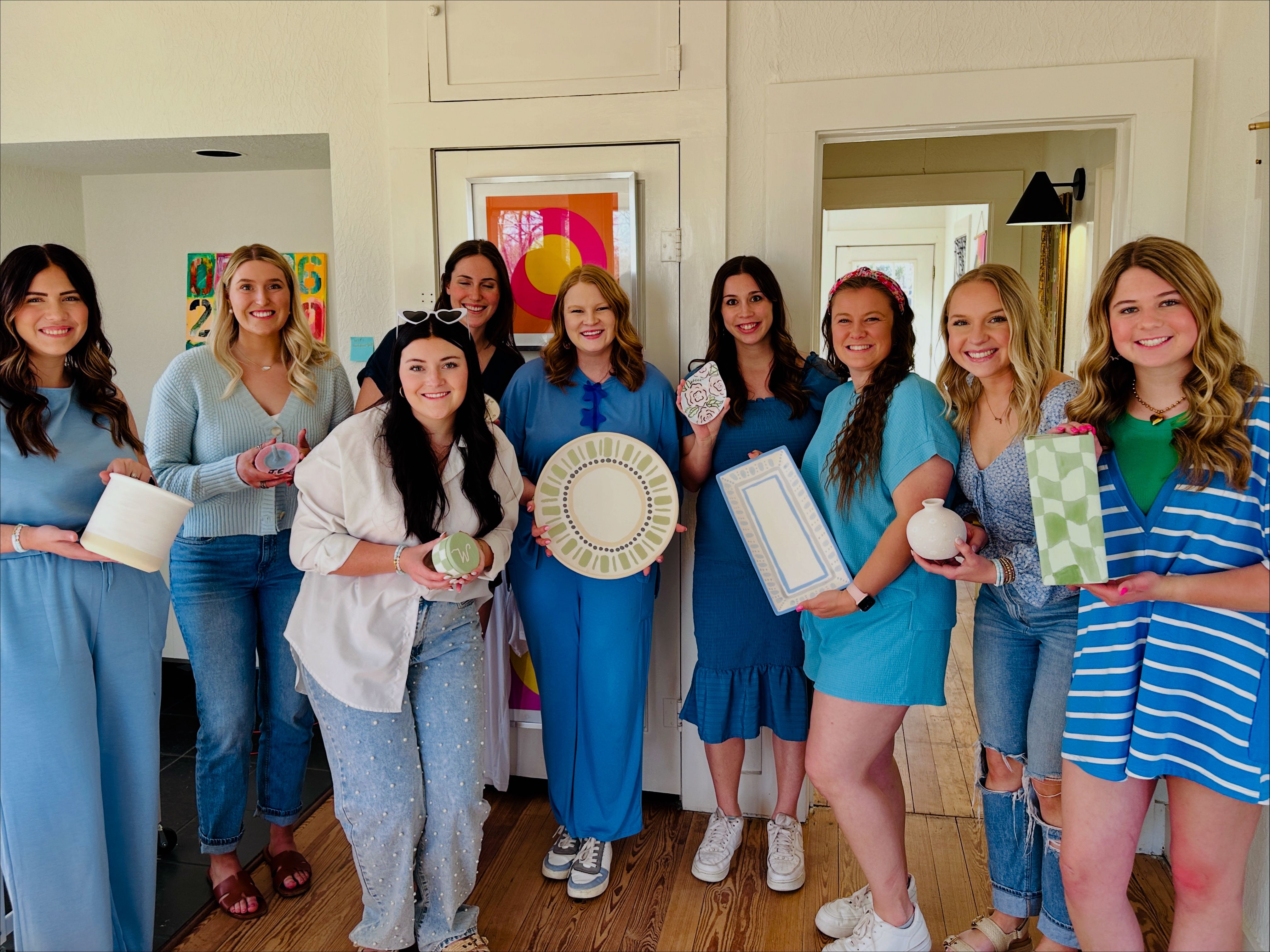 Smiling group of nine women wearing blue tones indoors holding handmade pottery, painted plates and gift-wrapped ceramics at a bright home DIY craft party