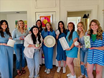 Smiling group of nine women wearing blue tones indoors holding handmade pottery, painted plates and gift-wrapped ceramics at a bright home DIY craft party