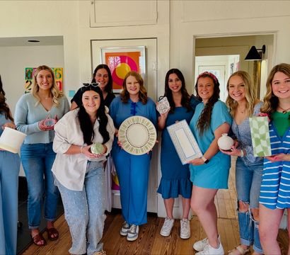 Smiling group of nine women wearing blue tones indoors holding handmade pottery, painted plates and gift-wrapped ceramics at a bright home DIY craft party