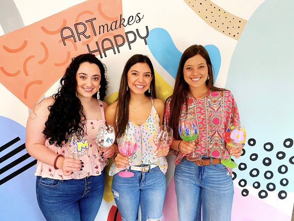 Three smiling women holding hand-painted wine glasses in a colorful paint-your-own-glass workshop, posed in front of a mural reading “ART makes HAPPY”.