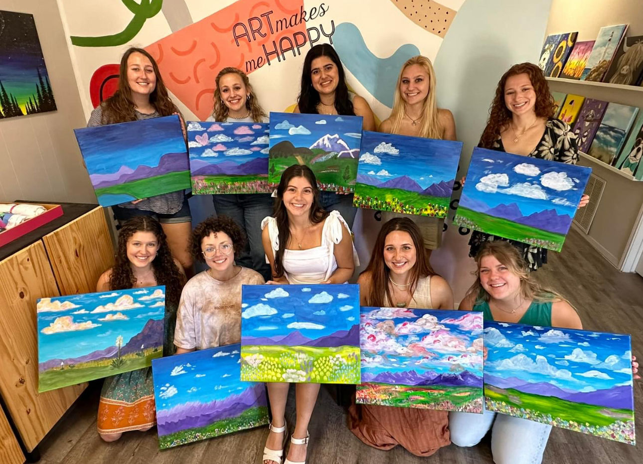 Painting class at a local art studio — a cheerful group holding colorful landscape canvases with blue skies, fluffy clouds, mountains and green fields.