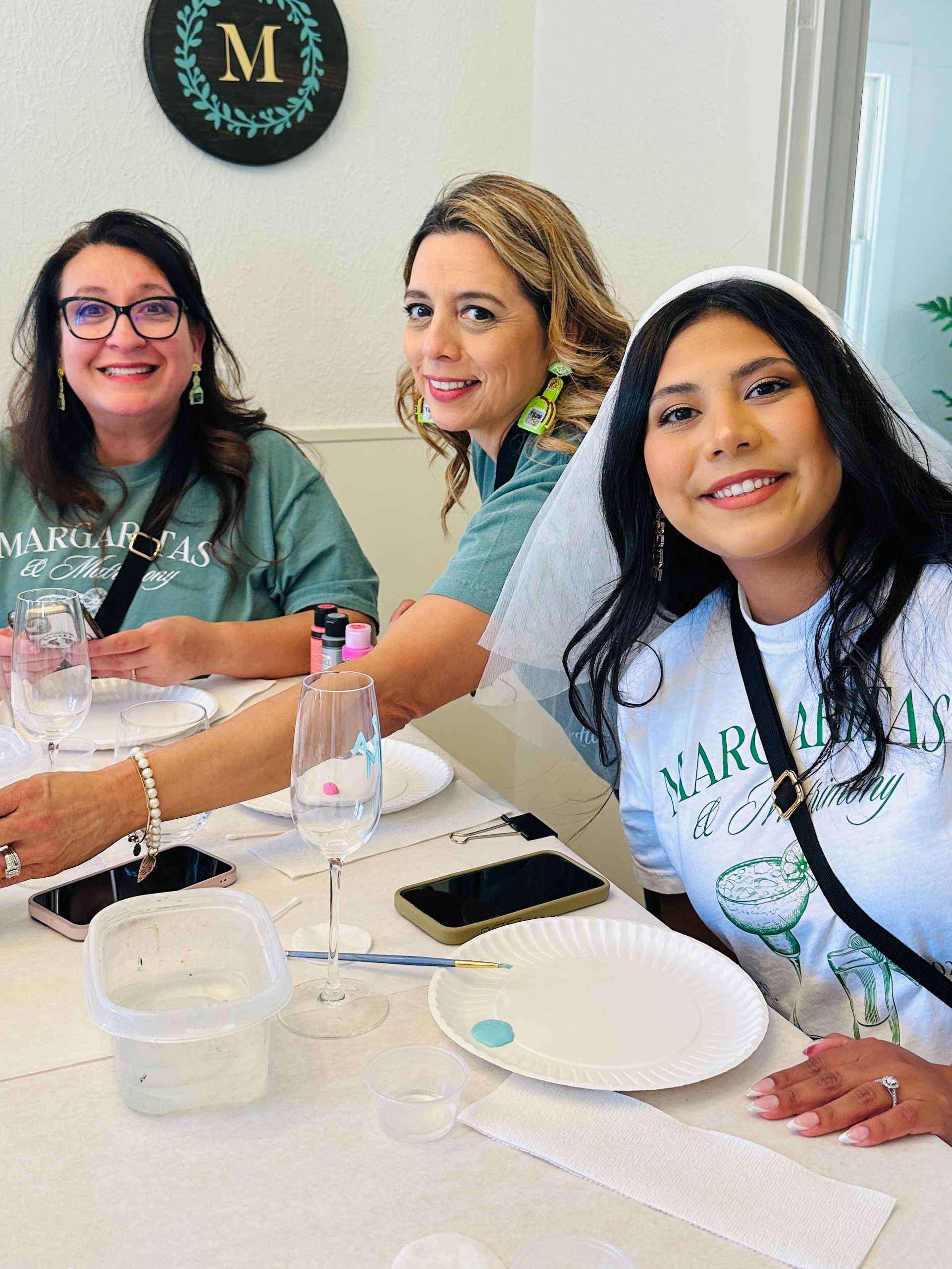 Three smiling women at an indoor margarita-themed bachelorette craft party — one wearing a white veil — seated at a table with wine glasses, paint, brushes and paper plates.