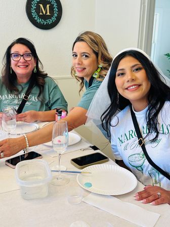 Three smiling women at an indoor margarita-themed bachelorette craft party — one wearing a white veil — seated at a table with wine glasses, paint, brushes and paper plates.