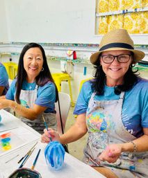 Two smiling women at a bright art studio workshop painting blue stripes on glassware, wearing paint-splattered aprons with brushes and palettes on the table, yellow stools and a patterned curtain in the background.