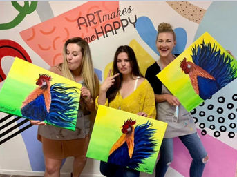 Three women at a lively art studio paint night holding colorful rooster canvas paintings in front of a mural that reads 'ART makes me HAPPY'.