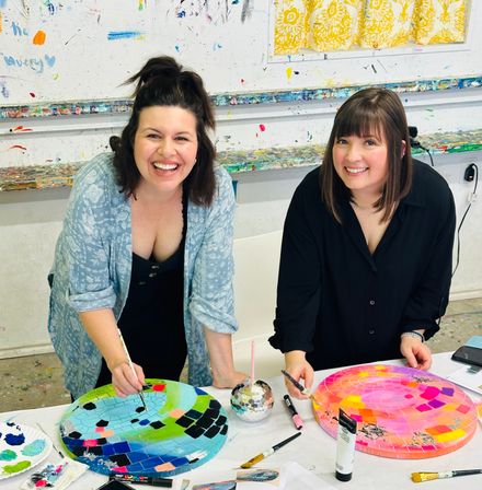 Two smiling women at a local art studio painting vibrant mosaic-style circular canvases on a paint-splattered table, brushes and colorful palettes nearby.