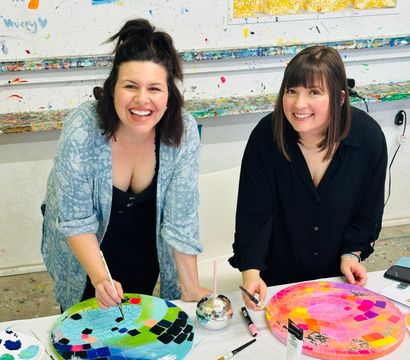 Two smiling women at a local art studio painting vibrant mosaic-style circular canvases on a paint-splattered table, brushes and colorful palettes nearby.
