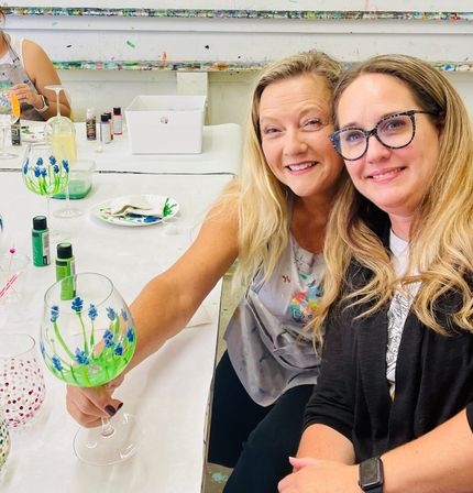 Two smiling women at a paint-and-sip workshop holding hand-painted wine glasses decorated with blue floral designs, seated at a white studio table with paint bottles and craft supplies