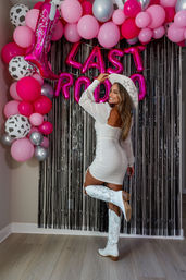 Woman in white mini dress, cowboy hat and white embroidered boots posing in front of pink and silver balloon arch and metallic fringe backdrop with hot pink letter balloons spelling 'LAST RODEO'