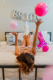 Woman lying on bed at a bachelorette celebration tossing pink balloons, wearing white cowboy boots and gingham dress with silver "BRIDE" balloons and a pink cowboy boot balloon in the background.
