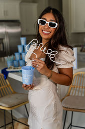Smiling woman in white sunglasses and cream dress holding a blue cup reading “Last toast on the coast” with a playful curly straw at a coastal-themed kitchen party
