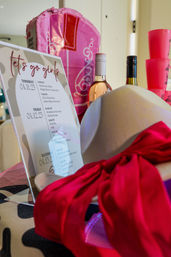 Bachelorette party welcome display with pink gift bag, printed itinerary sign, two wine bottles, stacked pink cups and a beige hat tied with a bright red scarf.