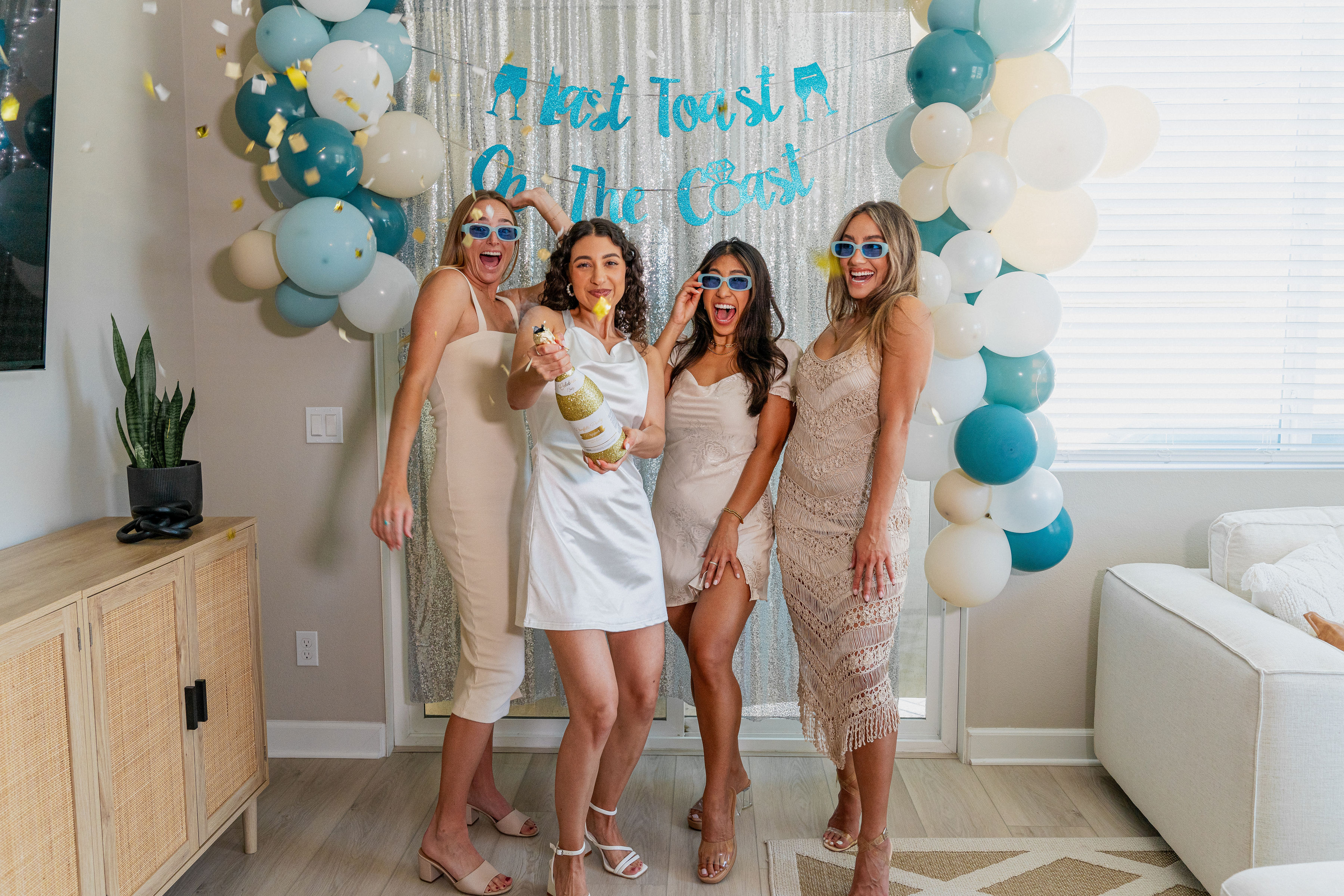 Four women in party dresses and blue sunglasses celebrate a coastal-themed bachelorette party indoors, the bride in white popping champagne in front of a silver sequin backdrop and teal-and-cream balloon arch.