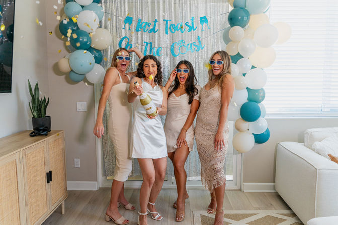 Four women in party dresses and blue sunglasses celebrate a coastal-themed bachelorette party indoors, the bride in white popping champagne in front of a silver sequin backdrop and teal-and-cream balloon arch.