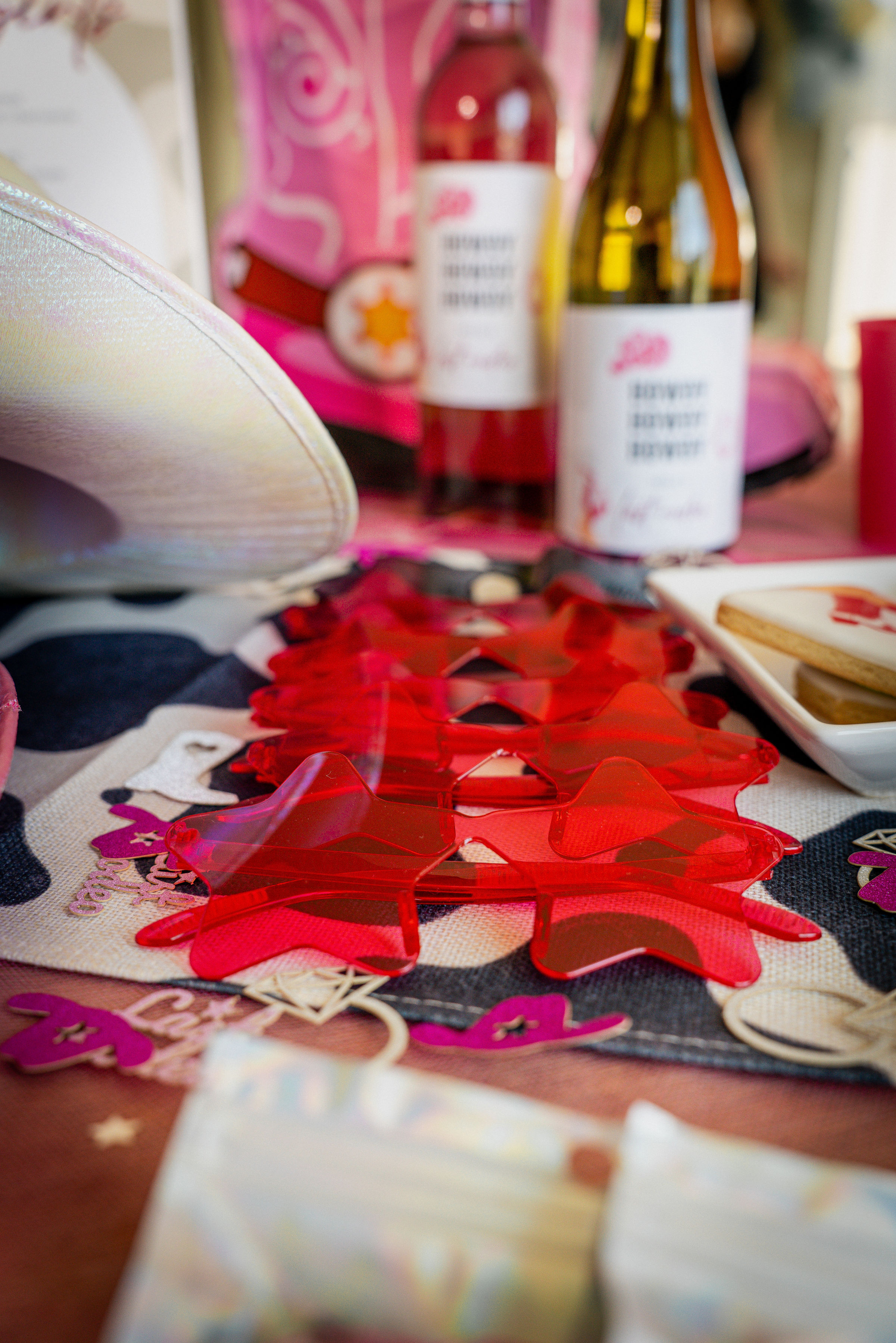 Bright red translucent star-shaped table confetti on a patterned runner with two wine bottles, a white cowboy hat and cookies in soft focus — pink-themed party display.