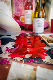 Bright red translucent star-shaped table confetti on a patterned runner with two wine bottles, a white cowboy hat and cookies in soft focus — pink-themed party display.