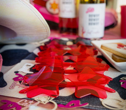 Bright red translucent star-shaped table confetti on a patterned runner with two wine bottles, a white cowboy hat and cookies in soft focus — pink-themed party display.