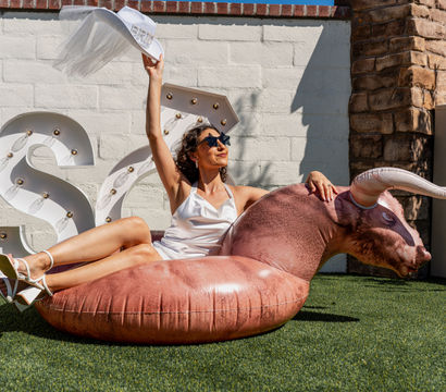 Woman in white dress and sunglasses lounging on a bull-shaped inflatable pool float on artificial turf in a sunny backyard patio, waving a hat near marquee light letters.