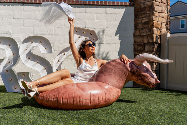Woman in white dress and sunglasses lounging on a bull-shaped inflatable pool float on artificial turf in a sunny backyard patio, waving a hat near marquee light letters.