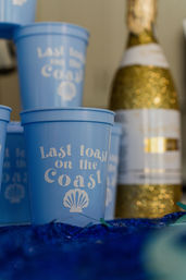 Blue plastic cup reading 'Last toast on the Coast' with a seashell graphic, stacked blue cups and a glittery gold champagne bottle blurred in the background, beach-themed celebration vibe.