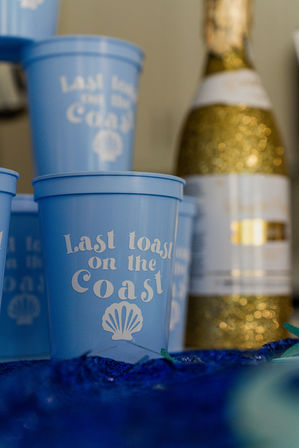 Blue plastic cup reading 'Last toast on the Coast' with a seashell graphic, stacked blue cups and a glittery gold champagne bottle blurred in the background, beach-themed celebration vibe.