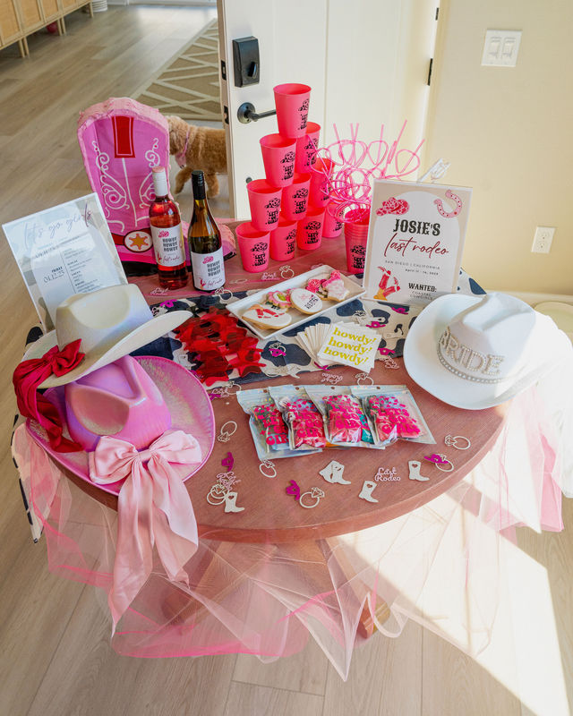 Bright pink cowgirl-themed bachelorette party table with white “BRIDE” hat, pink cowboy hats and bows, stacked pink cups and looped straws, wine bottles, decorated cookies, party favors and tulle table skirt.