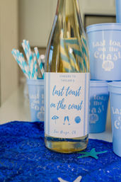 Blue-labeled wine bottle reading “last toast on the coast” on a sequined blue tablecloth, flanked by matching blue cups, striped paper straws and a starfish — San Diego, CA