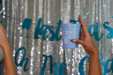 Raised hand holding a pastel blue cup with striped straw that reads 'Toast on the Coast' against a sparkly silver sequin backdrop at a coastal-themed party.