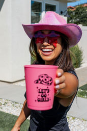 Smiling woman in a pink cowboy hat and star-shaped pink sunglasses holding a pink cup that reads "LET'S GO GIRLS" toward the camera at a sunny backyard gathering