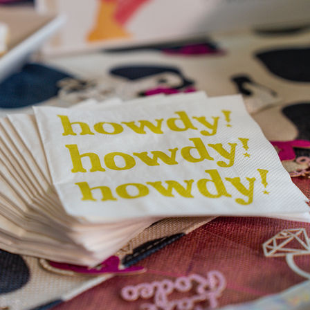 Stack of white paper napkins on a colorful table, each printed with repeating yellow "howdy!" text — playful western-style greeting
