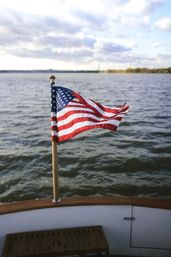 Small American flag on a wooden pole at a boat’s stern, fluttering proudly over rippling water with a distant shoreline and cloudy sky.