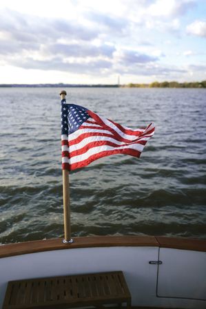 Small American flag on a wooden pole at a boat’s stern, fluttering proudly over rippling water with a distant shoreline and cloudy sky.