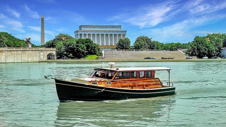 Classic wooden motorboat cruising the water in Washington, D.C., with the Lincoln Memorial and Washington Monument framed under a bright blue sky.