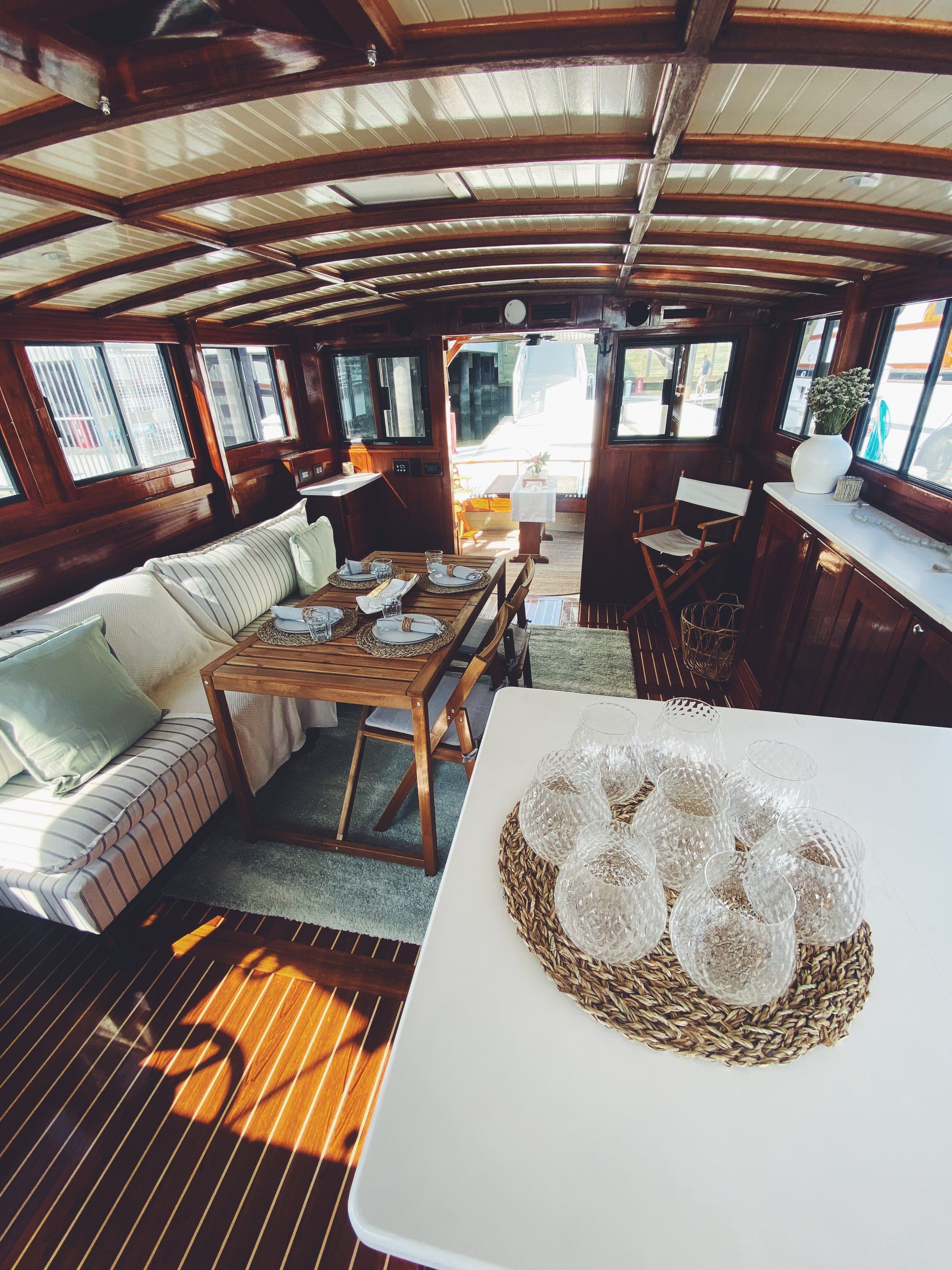 Cozy wood-paneled boat cabin interior with nautical dining area, foldable wooden table set for four, striped sofa, wicker placemat of glass tumblers, sunlight streaming through dockside windows