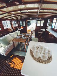 Cozy wood-paneled boat cabin interior with nautical dining area, foldable wooden table set for four, striped sofa, wicker placemat of glass tumblers, sunlight streaming through dockside windows