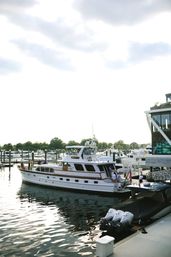 White luxury motor yacht docked at a busy marina waterfront, crew and guests boarding while smaller powerboats and docks reflect on rippling water