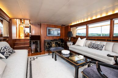 Sunlit luxury yacht salon interior with warm wood paneling, large windows framing coastal views, twin gray sofas with geometric pillows, black lacquer coffee table and decorative cabinet.