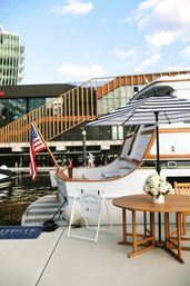 White yacht moored at a sunny marina dock with an American flag, black-and-white striped umbrella shading a wooden table topped with white hydrangeas, modern waterfront buildings in the background