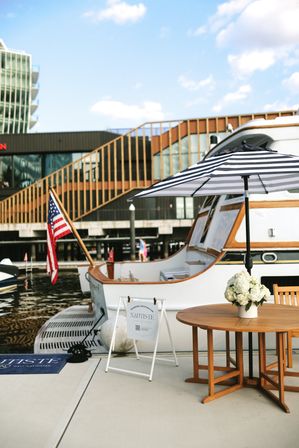 White yacht moored at a sunny marina dock with an American flag, black-and-white striped umbrella shading a wooden table topped with white hydrangeas, modern waterfront buildings in the background