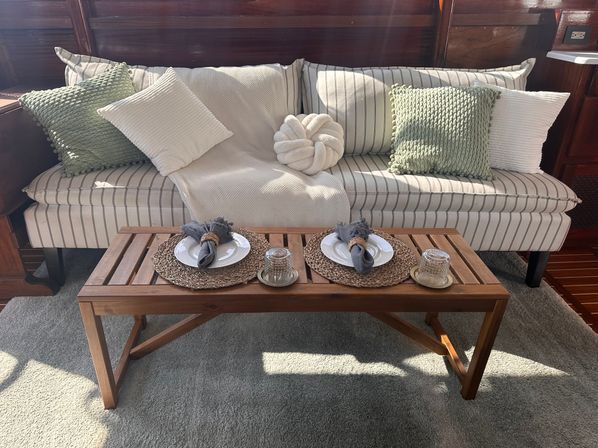 Sunlit cozy seating area: striped sofa with green and white textured pillows and a knotted cushion, cream throw, wooden slatted coffee table set for two with woven placemats and napkin-wrapped plates in a wood-paneled interior.