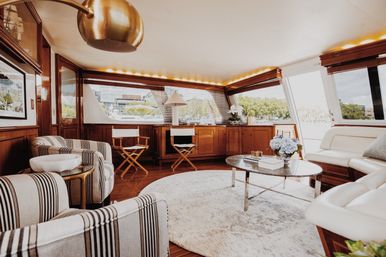 Sunlit luxury yacht interior with wood-paneled salon, striped armchairs, round rug, glass coffee table and marina waterfront view