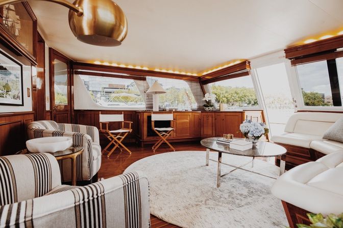 Sunlit luxury yacht interior with wood-paneled salon, striped armchairs, round rug, glass coffee table and marina waterfront view