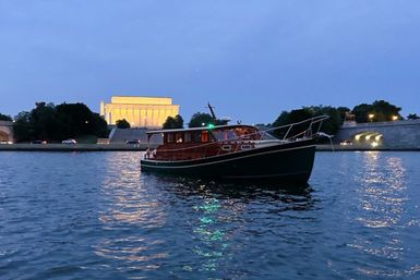 Cozy wooden cabin cruiser on the Potomac at dusk, with the illuminated Lincoln Memorial and riverfront bridge in Washington, D.C.