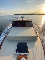 Bow of a classic wooden-cabin motorboat cruising calm open water at sunset, wake trailing toward a tree-lined shoreline with golden sun reflecting on the surface.