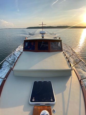 Bow of a classic wooden-cabin motorboat cruising calm open water at sunset, wake trailing toward a tree-lined shoreline with golden sun reflecting on the surface.