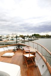 Wooden yacht deck with a folding canvas chair and bench, curved teak railing overlooking a marina of moored boats under a cloudy sky