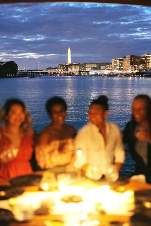 Candlelit riverside dinner at dusk with blurred guests in the foreground and the Washington Monument glowing above the illuminated Washington, D.C. waterfront