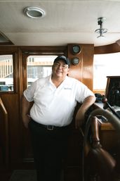 Cheerful boat captain in a white polo and cap leaning by a wooden helm inside a yacht cabin, with gauges on the wood panel and marina visible through the windows.